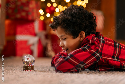 Happy Latin boy in red plaid shirt lying on the floor carpet, captivated by shimmering snow globe, side view