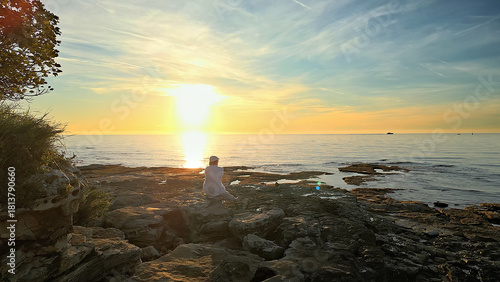 A solitary figure in white sits on rocky shores at sunset, facing the horizon. The scene evokes mindfulness and reflection, capturing a moment of solitude.