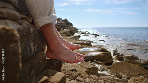 Fotografi A person leans against a rocky cliff, bare feet dangling over the edge, overlooking the ocean