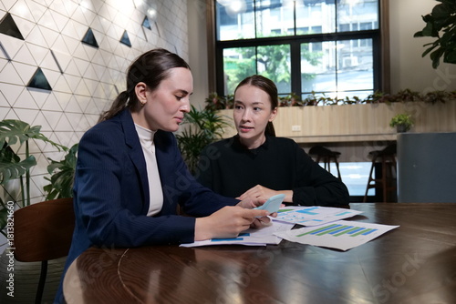 Two young Caucasian businesswomen discussing, review statistical data in paperwork and calculate business performance using a calculator in workplace.