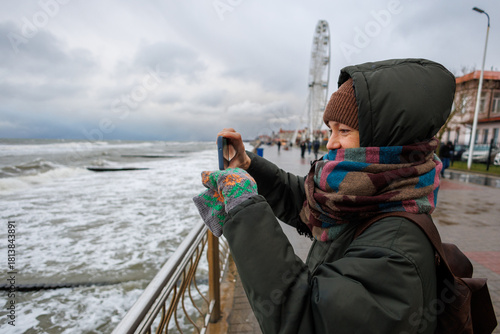 Person in winter clothing takes a photo by a stormy seaside. Overcast sky, Ferris wheel, and promenade in the background. Cold, windy atmosphere with waves crashing against the shore.