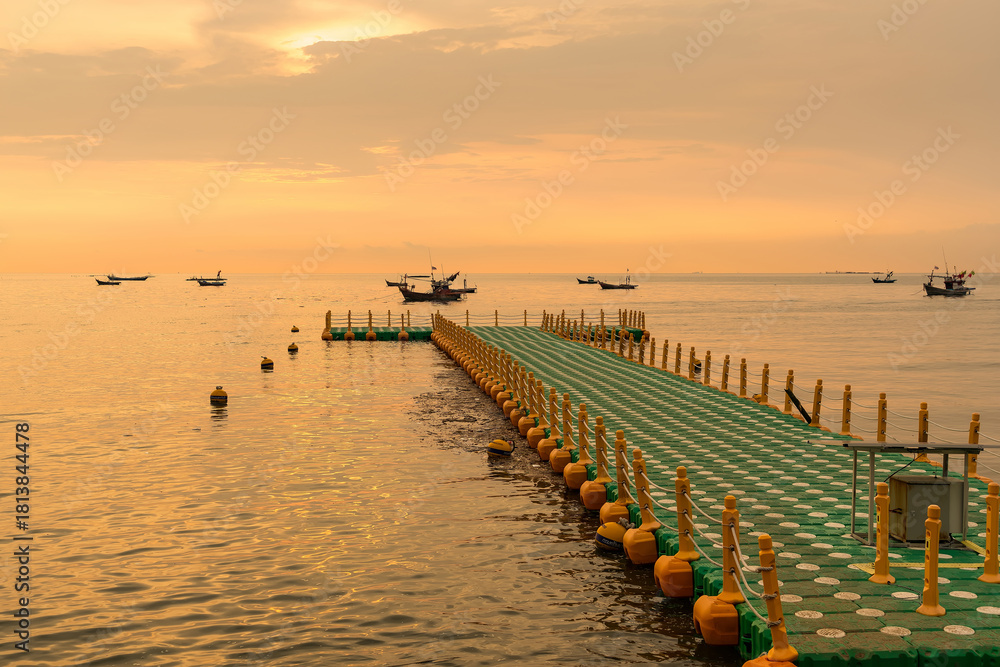 Obraz premium Floating dock extending into calm sea during golden sunrise with fishing boats on the horizon under a colorful sky