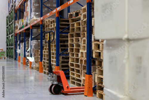 An orange manual pallet jack is parked on a concrete floor next to stacks of empty wooden pallets. The pallets are stored under tall blue and orange industrial racks in a modern warehouse.