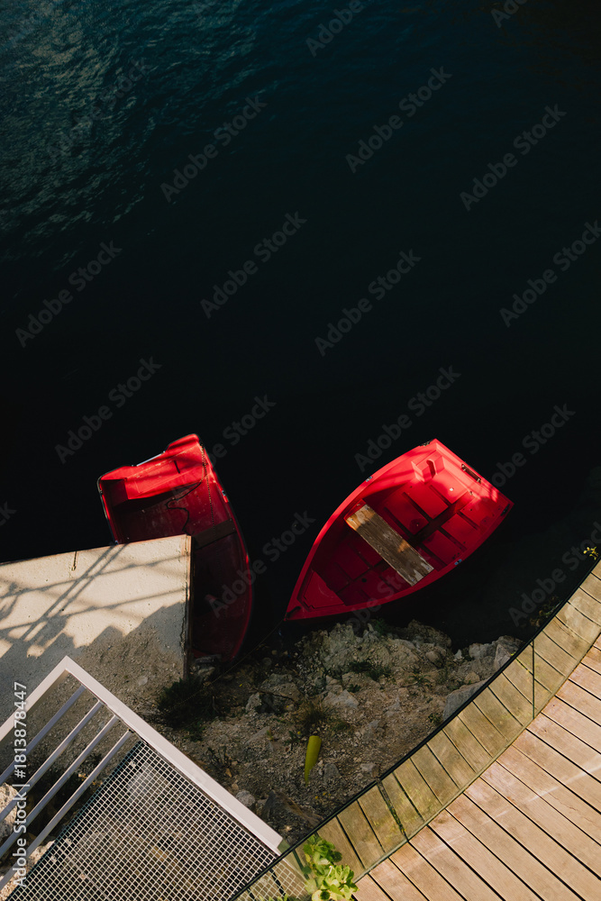 Fototapeta premium Two vibrant red boats sit near modern dock with wooden planks and concrete sections, creating striking contrast against dark, still water. Bright red boats mooring alongside sleek wooden concrete dock