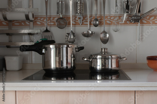 Stainless steel pressure cooker and cooking pot sitting on a modern induction hob with various kitchen tools hanging on the tiled wall in a domestic kitchen setting
