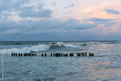 Fototapeta Naklejka Na Ścianę i Meble -  An evening seascape on the Baltic Sea. Low clouds and a shimmering sky. An old, worn log breakwater resists the breaking waves.