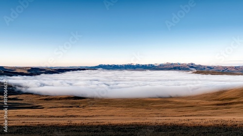 A panoramic view of a valley blanketed in dense clouds, with undulating golden hills in the foreground and majestic snow-capped mountains in the distance.