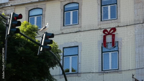 A seagull perched on a traffic light in front of a building decorated with Christmas lights and a large gift-shaped ornament.