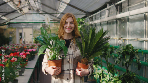 Woman holding potted plants in a greenhouse filled with colorful flowers during bright daylight hours