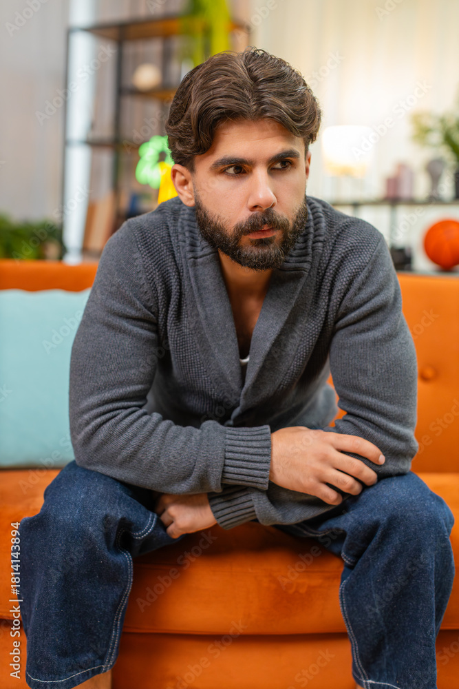 Fototapeta premium Indian man at home bends forward holds head in hands strong fatigue slow breath eyes down seeks rest. Arabian guy on sofa sits still rubs temples tries to calm mind seeks relief from stress and pain.