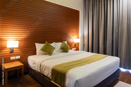 A bed with white sheets and green pillows sits against a wooden panel wall, with two wall lamps and small side tables creating a simple layout.