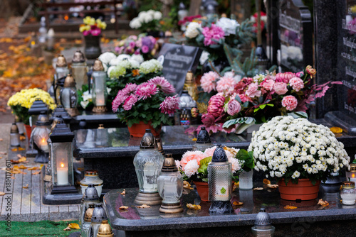 Zbliżenie na znicze i kwiaty zdobiące nagrobki na cmentarzu tuż po święcie zmarłych. Ciepłe światło zachodzącego słońca podkreśla spokojny i refleksyjny charakter miejsca, tworząc atmosferę zadumy i p