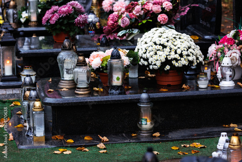 Zbliżenie na znicze i kwiaty zdobiące nagrobki na cmentarzu tuż po święcie zmarłych. Ciepłe światło zachodzącego słońca podkreśla spokojny i refleksyjny charakter miejsca, tworząc atmosferę zadumy i p