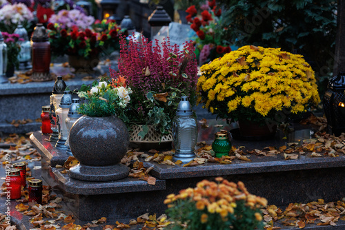 Zbliżenie na znicze i kwiaty zdobiące nagrobki na cmentarzu tuż po święcie zmarłych. Ciepłe światło zachodzącego słońca podkreśla spokojny i refleksyjny charakter miejsca, tworząc atmosferę zadumy i p