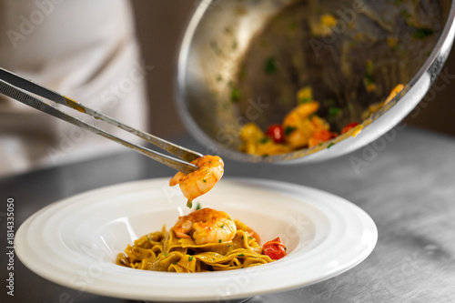 Fotografie A chef uses tongs to meticulously plate gourmet shrimp linguine pasta with cherry tomatoes and herbs in a professional kitchen setting, highlighting fine dining presentation and fresh ingredients