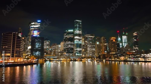 Wallpaper Mural Sydney city skyline at night, modern skyscrapers with illuminated windows and vibrant urban landscape, reflecting colorful lights on tranquil harbor waters during the evening. Drone flight footage Torontodigital.ca
