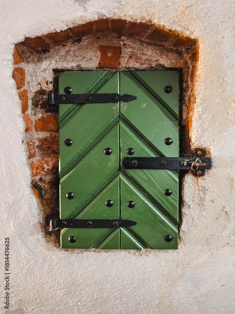 Obraz premium Small Green Shutter of Wooden Boards in the Shape of a Herringbone with Black Forged Hinges, Built into the Old Wall