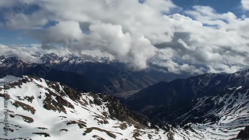 A panoramic aerial shot of snow-capped mountain peaks under a cloudy sky. The foreground shows snowy ridges with deep valleys