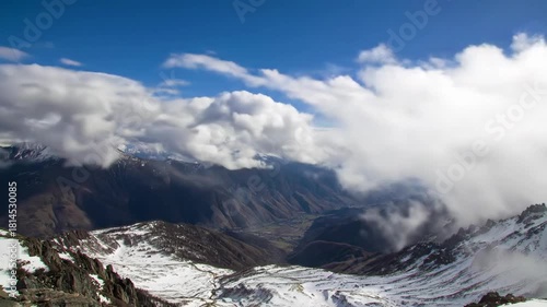 A panoramic view of a mountain range. The vast landscape includes snow-covered peaks, valleys, and clouds against a blue sky