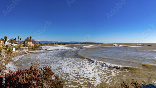 Fototapeta Naklejka Na Ścianę i Meble -  Sunny Mediterranean coast with brown stormy water, bright blue sky, foamy waves, traditional houses and snowy mountains along a quiet shoreline. Side, Turkey.