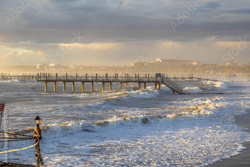 Fototapeta Naklejka Na Ścianę i Meble -  A surreal storm scene under bright sunlight: powerful waves flood the beach beyond limits, turning a ramp into an abyss. Nature's wild triumph in November. Side, Antalya, Turkey, Mediterranean.

