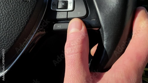 Switching music track using steering wheel control buttons inside modern car interior with close-up view of driver hand operating multimedia system