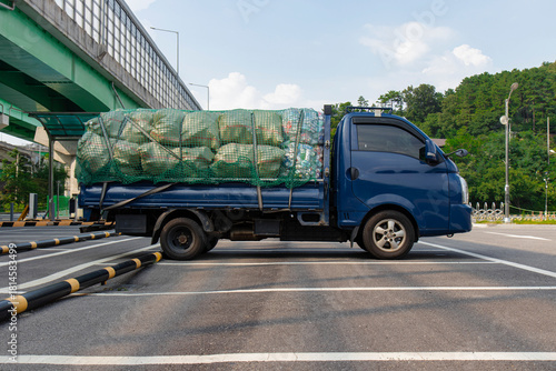 Fototapeta Naklejka Na Ścianę i Meble -  A small blue truck parked outdoors, filled with sacks covered with a green net.