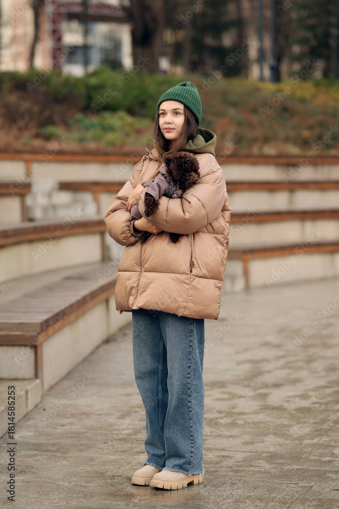 Fototapeta premium Young woman holding small poodle dog outdoors in autumn park, showing pet care, companionship and everyday lifestyle.