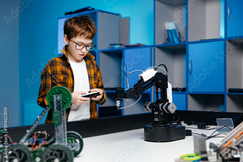 Young boy with glasses is programming a robotic arm in a modern classroom, surrounded by various robotics kits and tools, showcasing hands-on learning and innovation