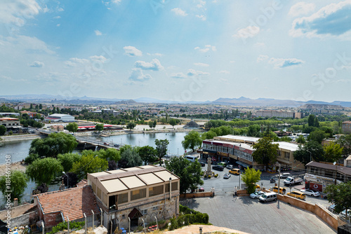 Fototapeta Naklejka Na Ścianę i Meble -  A vibrant urban landscape featuring a riverside area with green trees, buildings, bridges, and distant mountains under a bright, partly cloudy sky.
