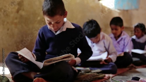 Raw footage of Indian village school boy reading textbook while sitting on classroom floor with other students inside rural government school in India.