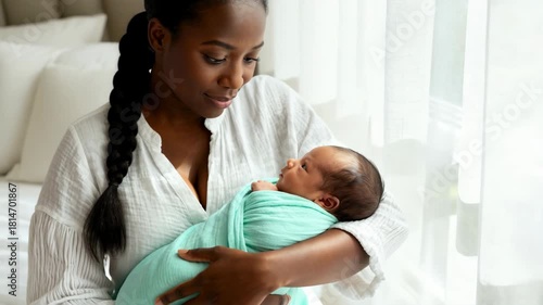 African American mother holding her newborn baby wrapped in a teal swaddle near a window. Mom looking lovingly at her infant child in a bright bedroom. Motherhood and family bonding concept