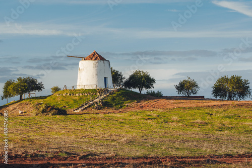 white windmill with a tiled roof sits peacefully on a green hill surrounded by scattered trees and farmland.