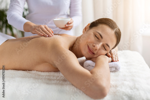 Young woman enjoying back massage while therapist applies exfoliating salt. Warm tones, gentle focus and soft texture highlight beauty care, skincare and deeply soothing spa treatment experience