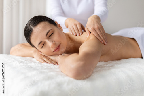 Young female smiling softly while receiving relaxing shoulder massage at spa. Warm light and gentle atmosphere emphasize comfort, beauty care, skincare and a joyful wellness lifestyle moment