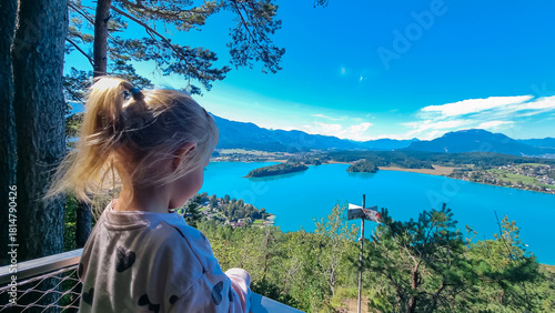 A young girl with blonde hair, seen from behind, stands at a scenic viewpoint and admires the breathtaking panoramic view of the turquoise Lake Faak and majestic Austrian Alps on beautiful sunny day