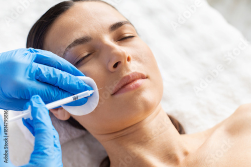 Young woman receiving cheek area aesthetic injection using cotton pad support. Clean spa setting and precise gloved technique highlight cosmetic care, beauty enhancement and dermatology precision