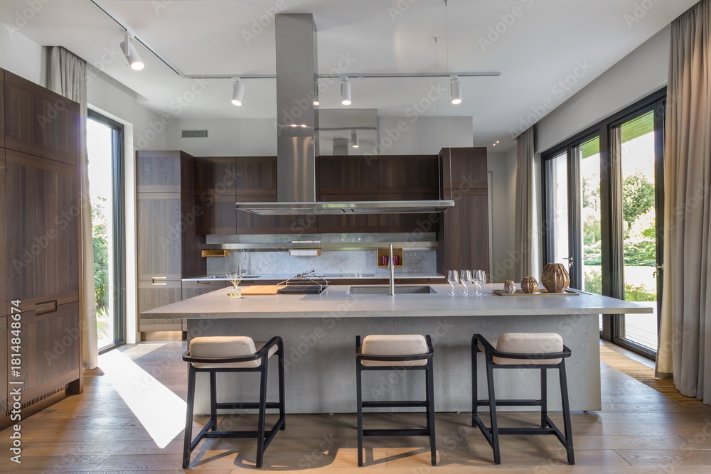 Fototapeta premium This stylish kitchen features a large island with three bar stools, modern cabinets, and windows that provide plenty of light. The open space showcases wood accents and is perfect for cooking