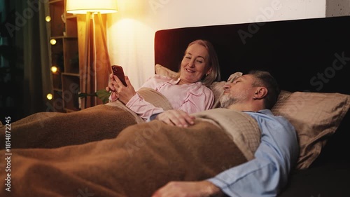 Middle-aged woman texting on phone next to sleeping husband, using dating app