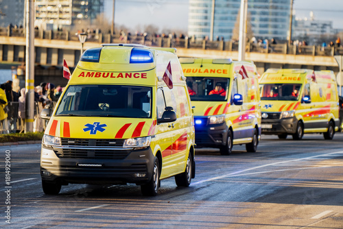 Yellow ambulances with blue emergency lights and Latvian flags moving in convoy along city roadway with medical symbols and red striping visible near bridge and crowd