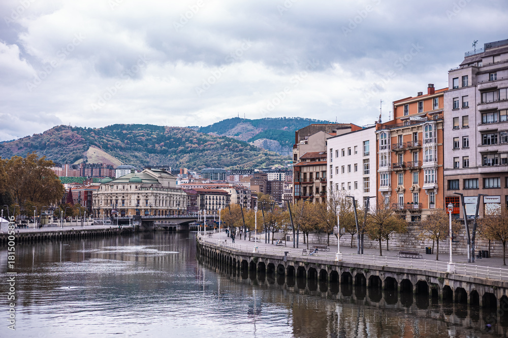 Fototapeta premium wide view of the Bilbao riverfront, with colorful buildings reflected in the calm water. The city rises up a wooded hillside under a dramatic, cloudy sky, showcasing urban and natural elements.