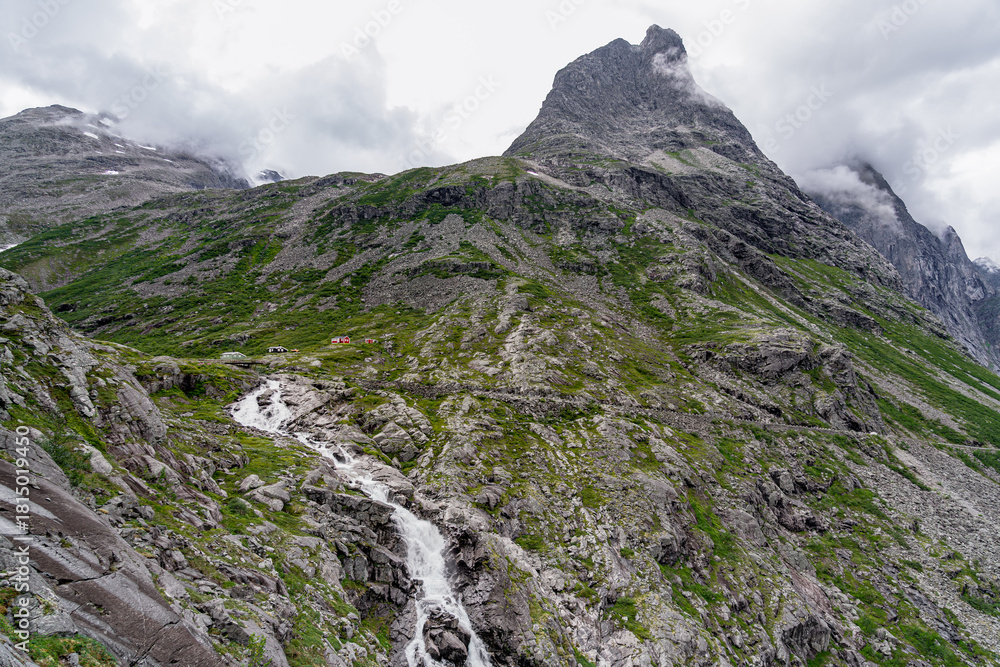 Obraz premium Remote houses at de foot of the Bispen mountain towers above the lush rocky landscape, with the Stigfossen waterfall cascading down its rocky slopes near the scenic Trollstigen road in Norway