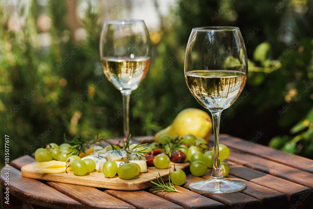 Fototapeta premium Two wine glasses with white wine and cheese board served with grapes and pear on wooden table outdoors