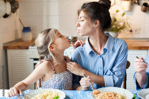 Mother and daughter eating pasta together at home kitchen. Family having fun, sharing spaghetti kiss. Kid enjoying homemade dinner, healthy comfort food. Happy childhood. Candid, authentic lifestyle