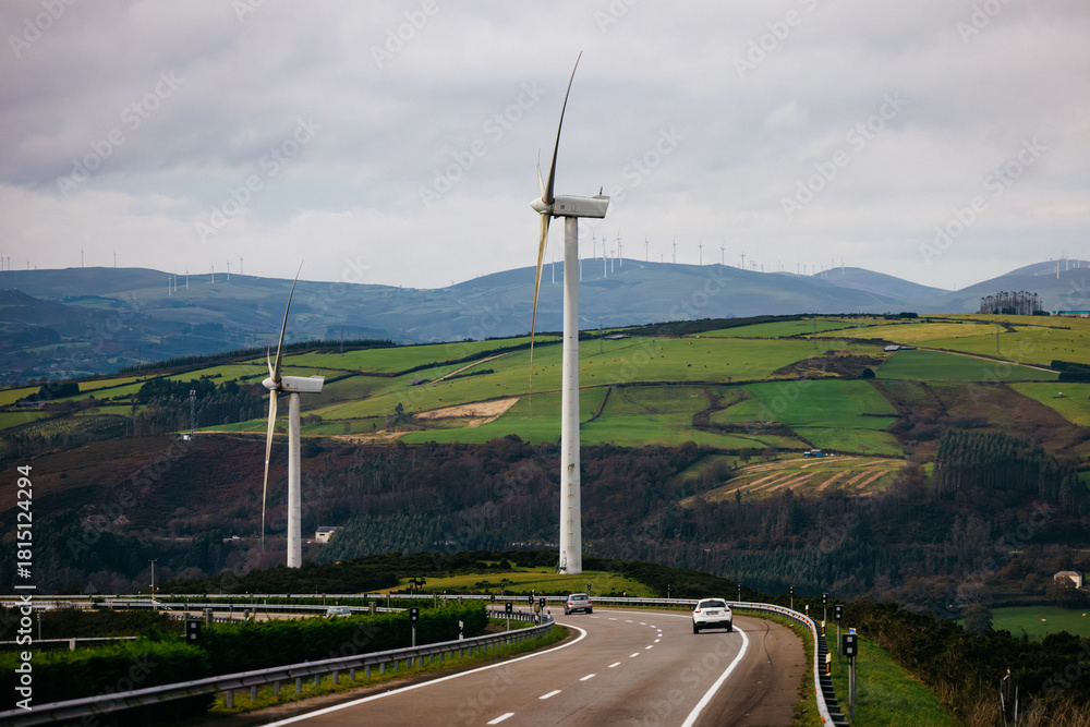 Naklejka premium Wind turbines stand tall on rolling green hills beside a winding road, showcasing renewable energy and sustainable technology in a serene landscape