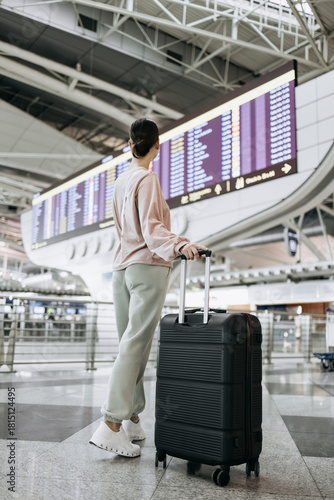 Young woman with a suitcase stands in an airport terminal, gazing at the flight information board, surrounded by modern architecture and a bustling travel atmosphere