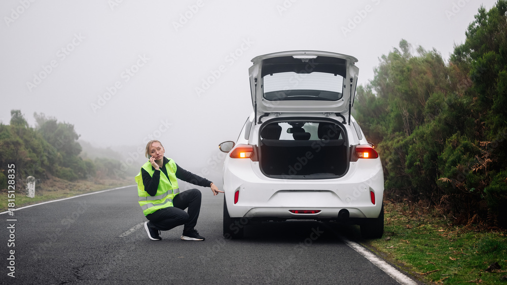 Obraz premium A woman wearing a reflective vest kneels next to a broken down car on a foggy road, demonstrating emergency repair and assistance.