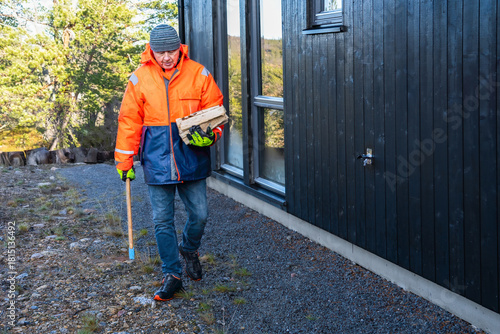 Steady practical movement as a man carries firewood and an axe toward a dark cabin, preparing for heating tasks with clear intent in bright Nordic daylight. Warm textures and Copy space.