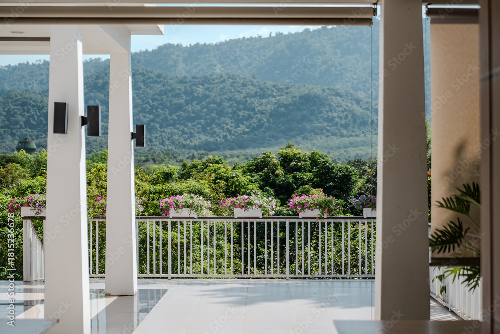 Fototapeta premium A bright terrace overlooking green hills features rows of colorful flowers along the railing, with clean white pillars framing the open space and highlighting the mountain scenery.