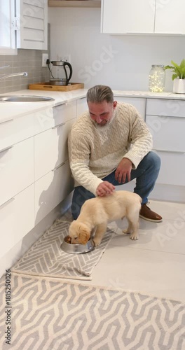Vertical video: Man placing dog bowl in kitchen causing puppy eating kibble, petting it, copy space